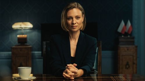 „Różyczka 2” do 10 listopada za darmo w TVP VOD. Magdalena Boczarska w eleganckim, ciemnym garniturze siedzi przy stole w gabinecie z polskimi flagami w tle. Patrzy prosto w obiektyw z poważnym, skupionym wyrazem twarzy.