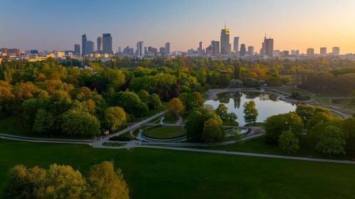 Widok z lotu ptaka na park z jeziorkiem oraz inne zielone tereny na obrzeżach Warszawy o zachodzie słońca.