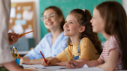 Uśmiechnięte dziewczynki siedzą w ławce szkolnej i słuchają nauczycielki. 